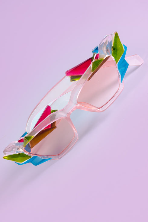 A pair of geometric sunglasses with angular frames featuring vibrant green, blue, and pink accents. The lenses are tinted light pink, adding a playful, modern twist to the bold, colorful design. These sunglasses make a striking, artistic statement